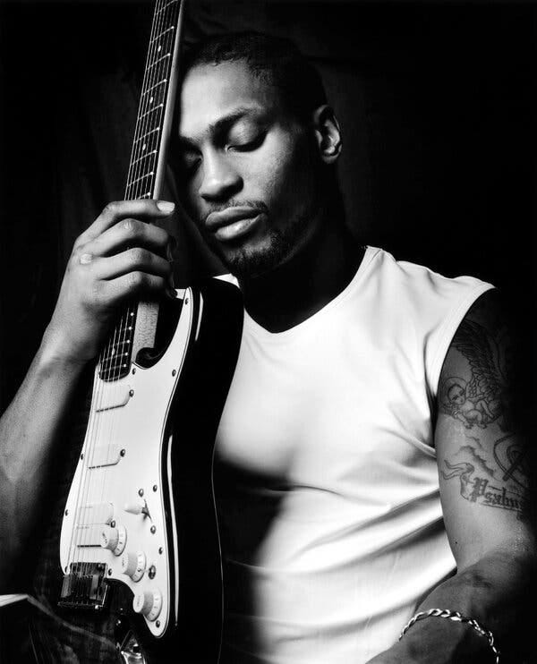 A black-and-white image of a man with his eyes closed, wearing a white sleeveless shirt with a tattoo on his arm and holding a guitar.