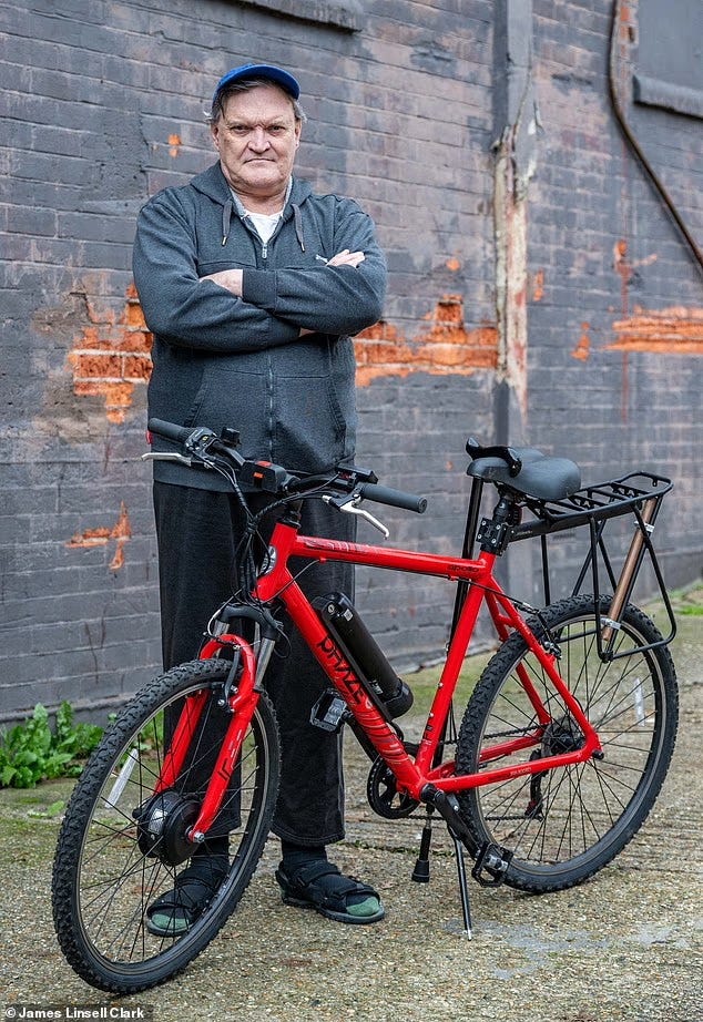 Robert Brown, 59, standing with his arms crossed beside a red bicycle against a brick wall in Colchester, Essex. He is fully clothed, wearing a dark hoodie and cap, and was photographed after being assaulted during a World Naked Bike Ride event.