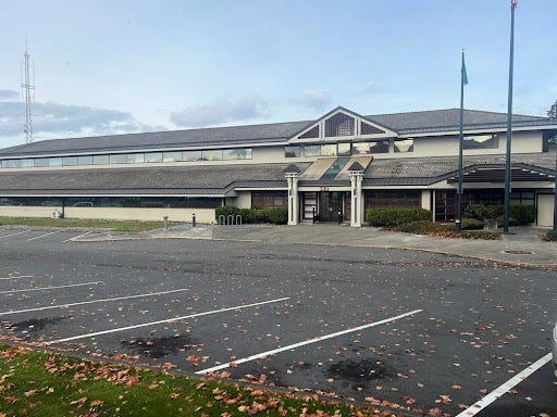 A side-angle photograph taken from the parking lot, showing the modern, two-story Port Angeles City Hall and Police Department building. The building features light cream-colored siding, extensive windows, a dark shingled roof with several distinct gables, and flagpoles (displaying the US and Washington State flags) next to the entrance.