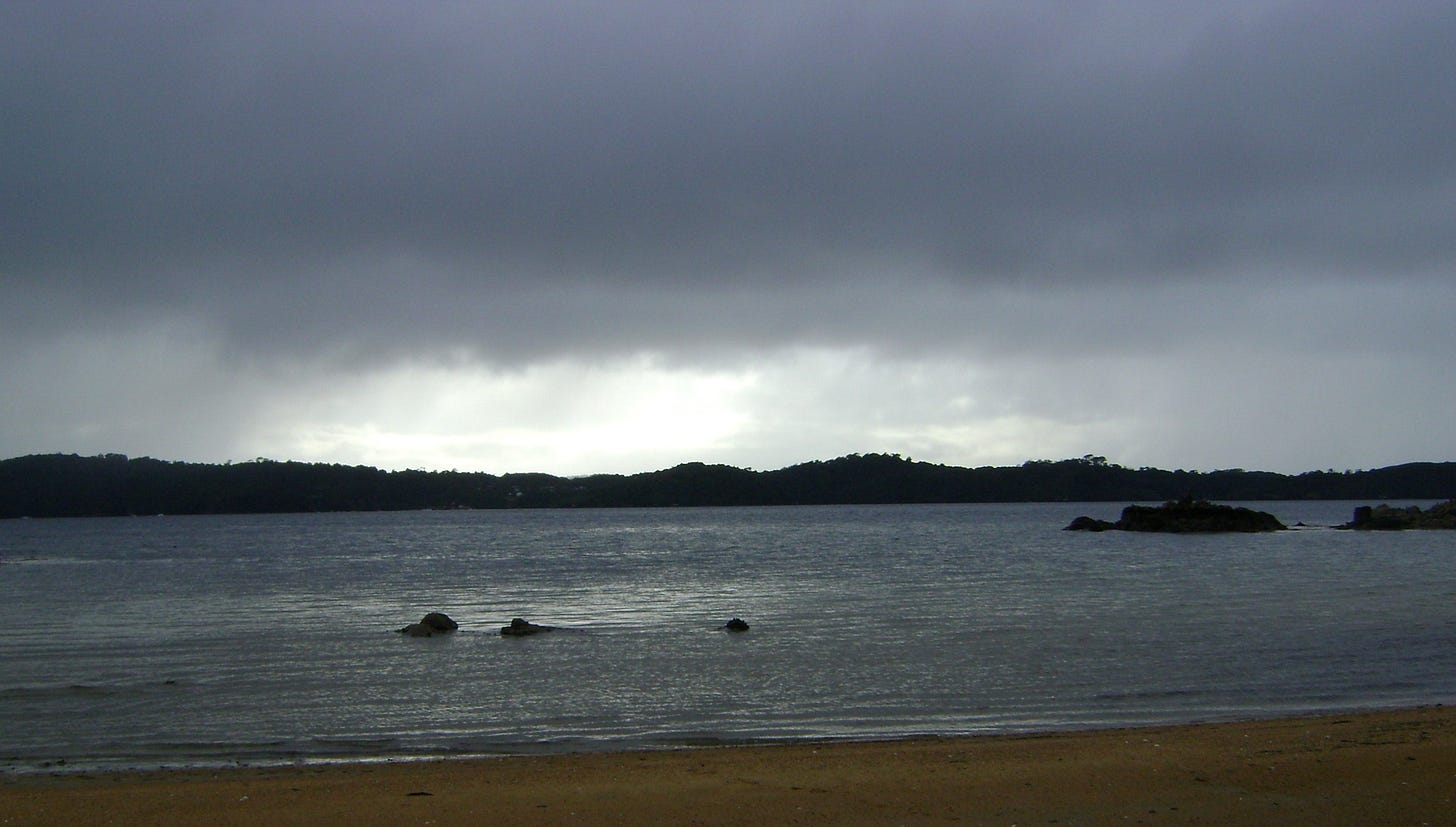 Image is of a beach with dark clouds, but a source of bright light between the sky and the see. Image is of a beach with dark clouds, but a source of bright light between the sky and the see.