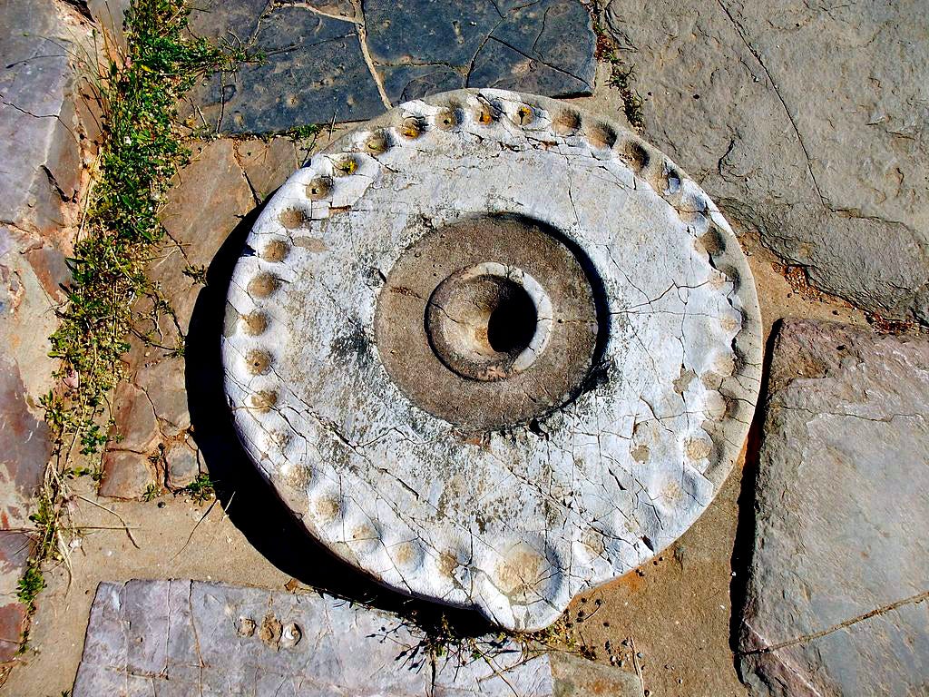 Circular stone kernos from Malia with a larger central cup-shaped depression and multiple smaller ones in a circle around the edge, punctuated with one slightly larger on toward the bottom of the photo. The kernos sits on an outdoor surface of rough stone paving.