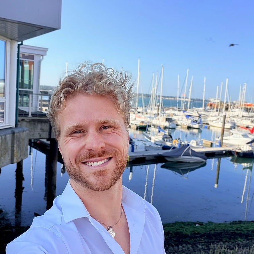 James S. Bayley smiling beside a marina filled with sailboats on a clear day. James S. Bayley smiling beside a marina filled with sailboats on a clear day.