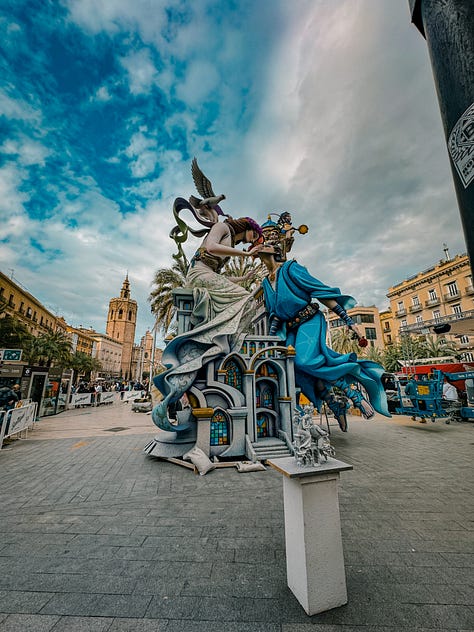 A gallery of Fallas monuments in Valencia, Spain — elaborately sculpted figures including a Charlie Chaplin figure holding butterflies, fantasy warriors, and colorful street sculptures, photographed against dramatic cloudy and blue skies in March 2026.