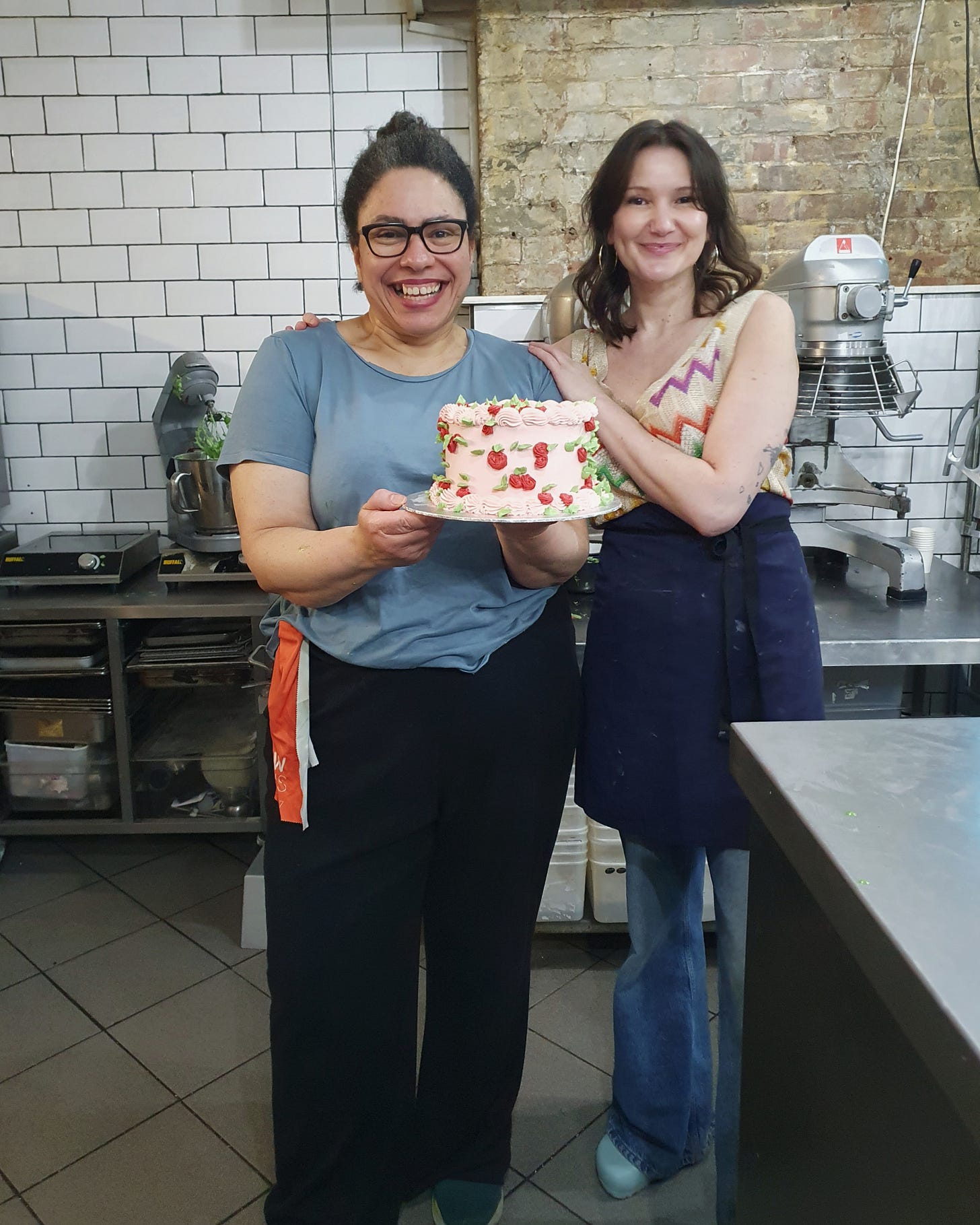 Photo of two women smiling to the camera, one has her arms around the other and that one is holding an iced cake.
