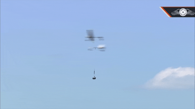 Two small unmanned aircraft fly across a clear blue sky while one lowers a small cylindrical payload suspended on a cable. Two small unmanned aircraft fly across a clear blue sky while one lowers a small cylindrical payload suspended on a cable.