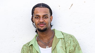 Callum wears an open green shirt over a white vest with a silver necklace and a chain