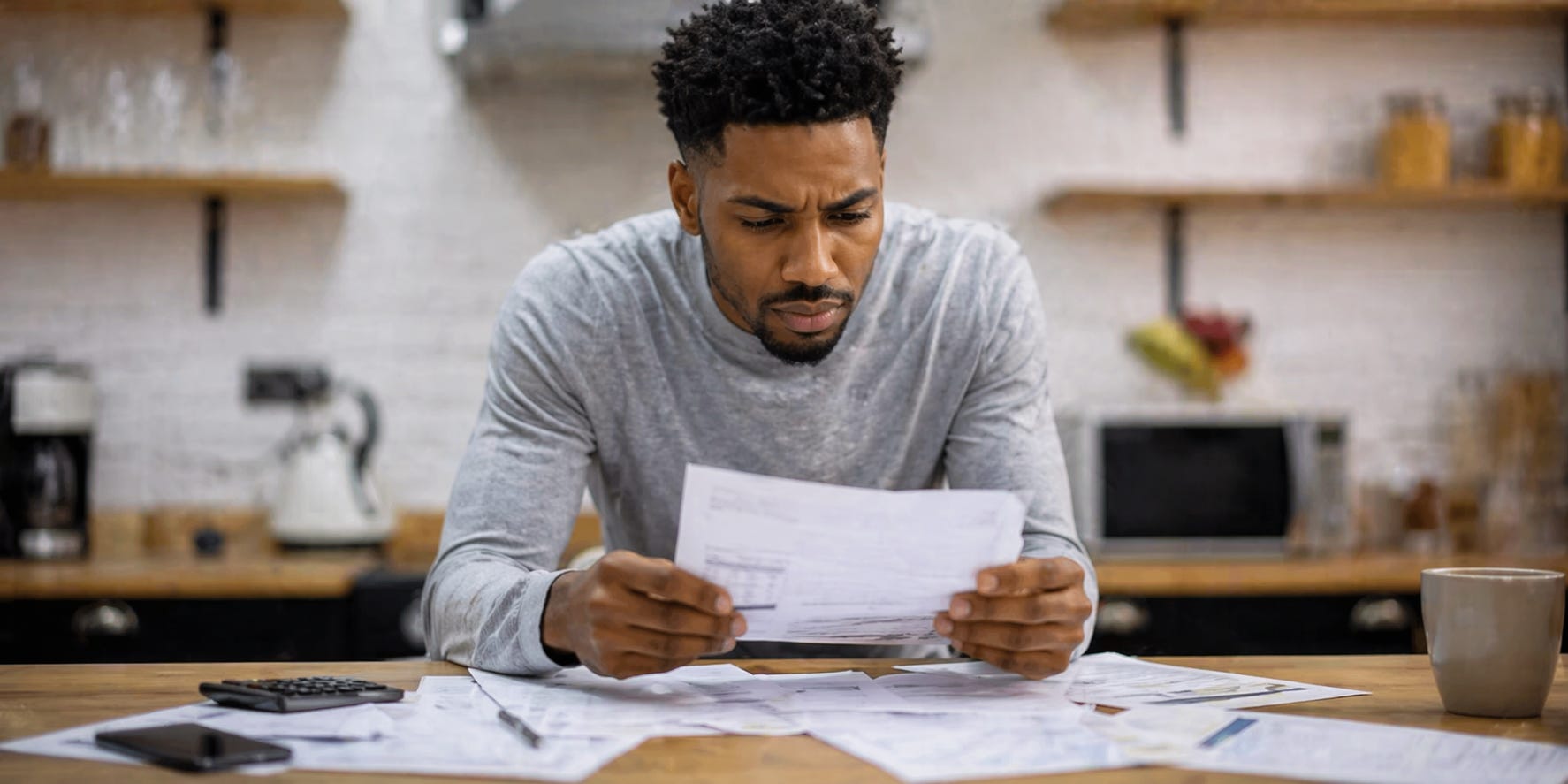 A man sits at a table covered in papers, studying a document with a concerned, questioning expression. The softly blurred background draws focus to the tension of managing finances and unexpected costs.