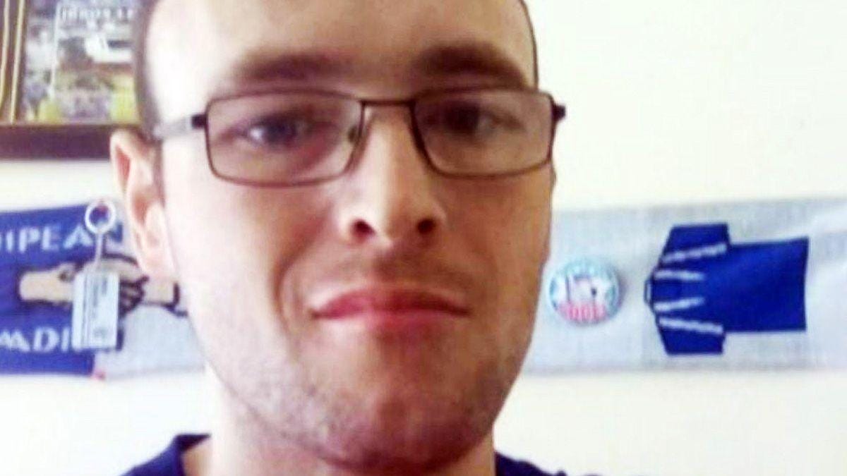 A man with short brown hair and glasses looks at the camera. He appears to be in a bedroom or living room