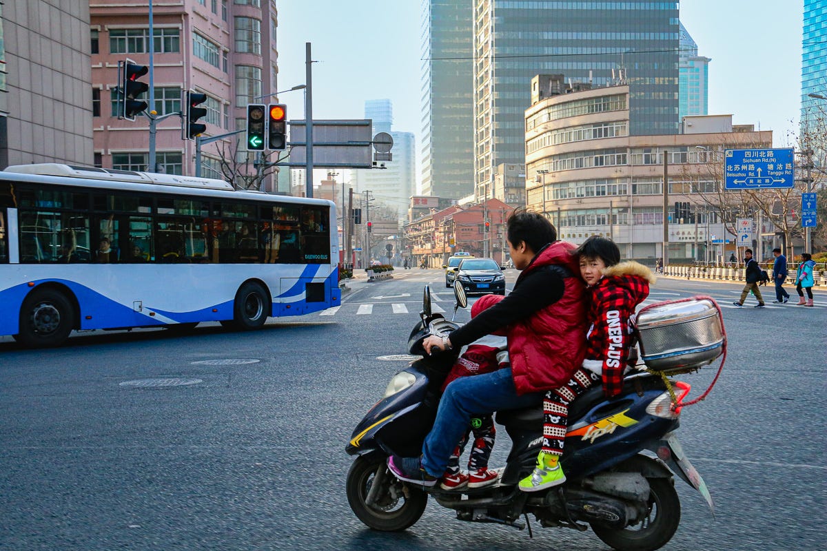 A man and two children ride a moped across a bustling Shanghai intersection. Modern high-rises and a city bus frame the scene of urban movement.