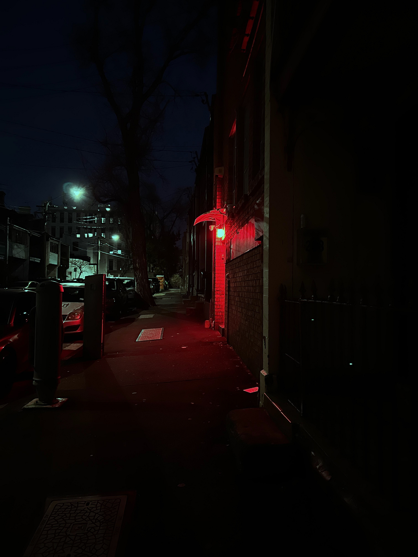 A dark, nighttime street scene illuminated by a red light on a building's exterior. The red light casts a glow on the pavement and nearby wall, while the rest of the street is dimly lit by distant streetlights and moonlight. The street is lined with parked cars and trees, with buildings visible in the background. The overall atmosphere is moody and mysterious