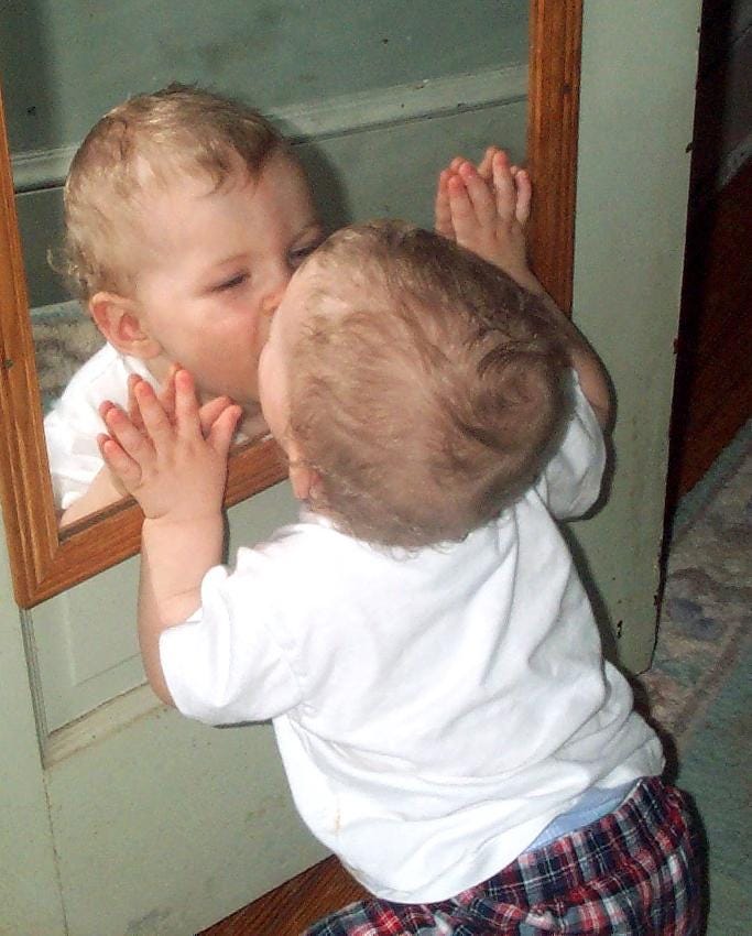 a baby squatting down next to a wall mirror and kissing it; baby has blonde hair and a white shirt a baby squatting down next to a wall mirror and kissing it; baby has blonde hair and a white shirt