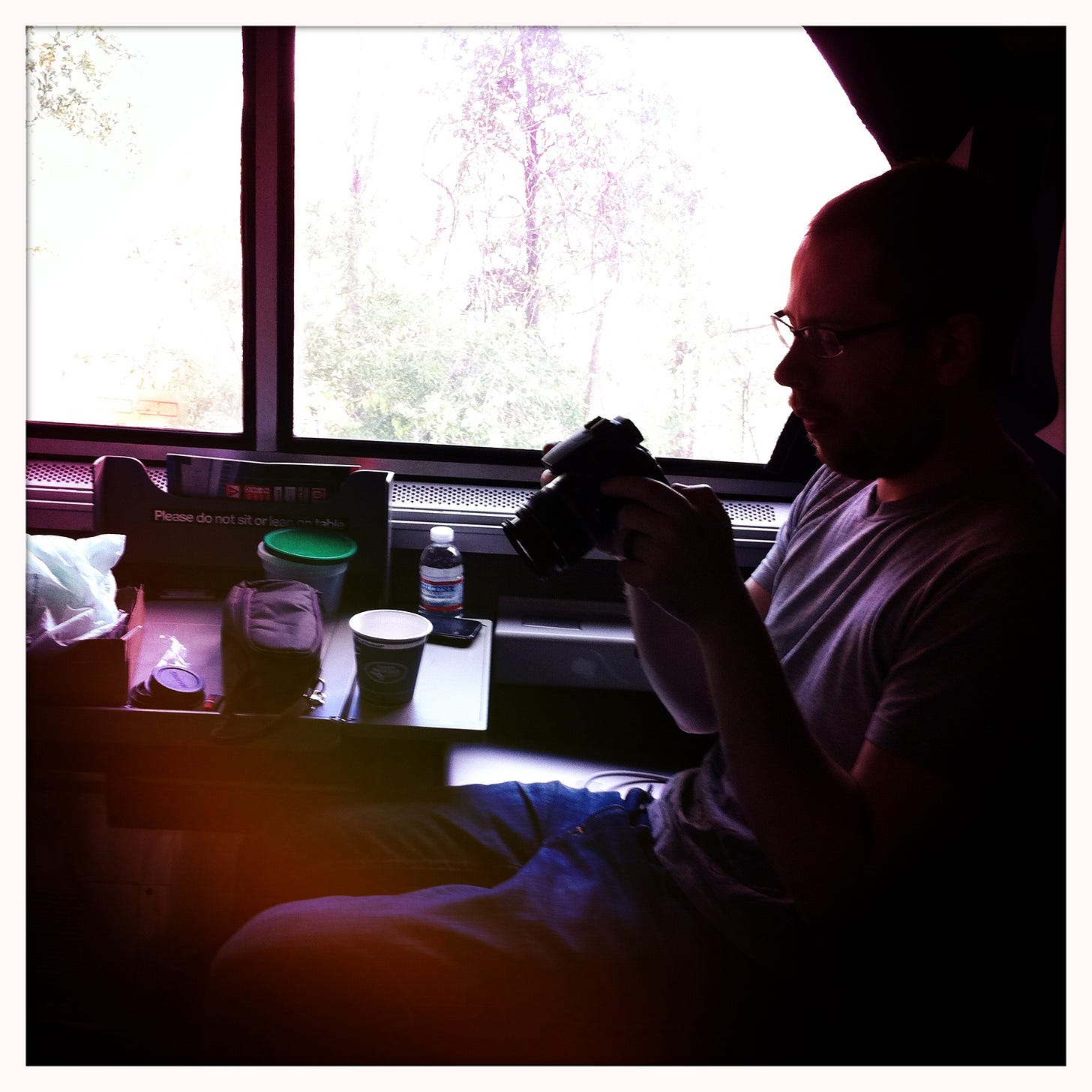 A man with glasses sits by a train window, holding a DSLR camera and looking down at it. He's in profile, wearing a grey t-shirt and jeans. The window behind him is blown out with bright daylight, showing the silhouettes of trees outside. On the table in front of him: a takeaway coffee cup, a water bottle, a small grey pouch, and a green-lidded container, with a "Please do not sit or lean on table" sign visible on the partition. The image has a heavy lo-fi filter aesthetic — vignetting, light leaks in pink and orange across the frame.