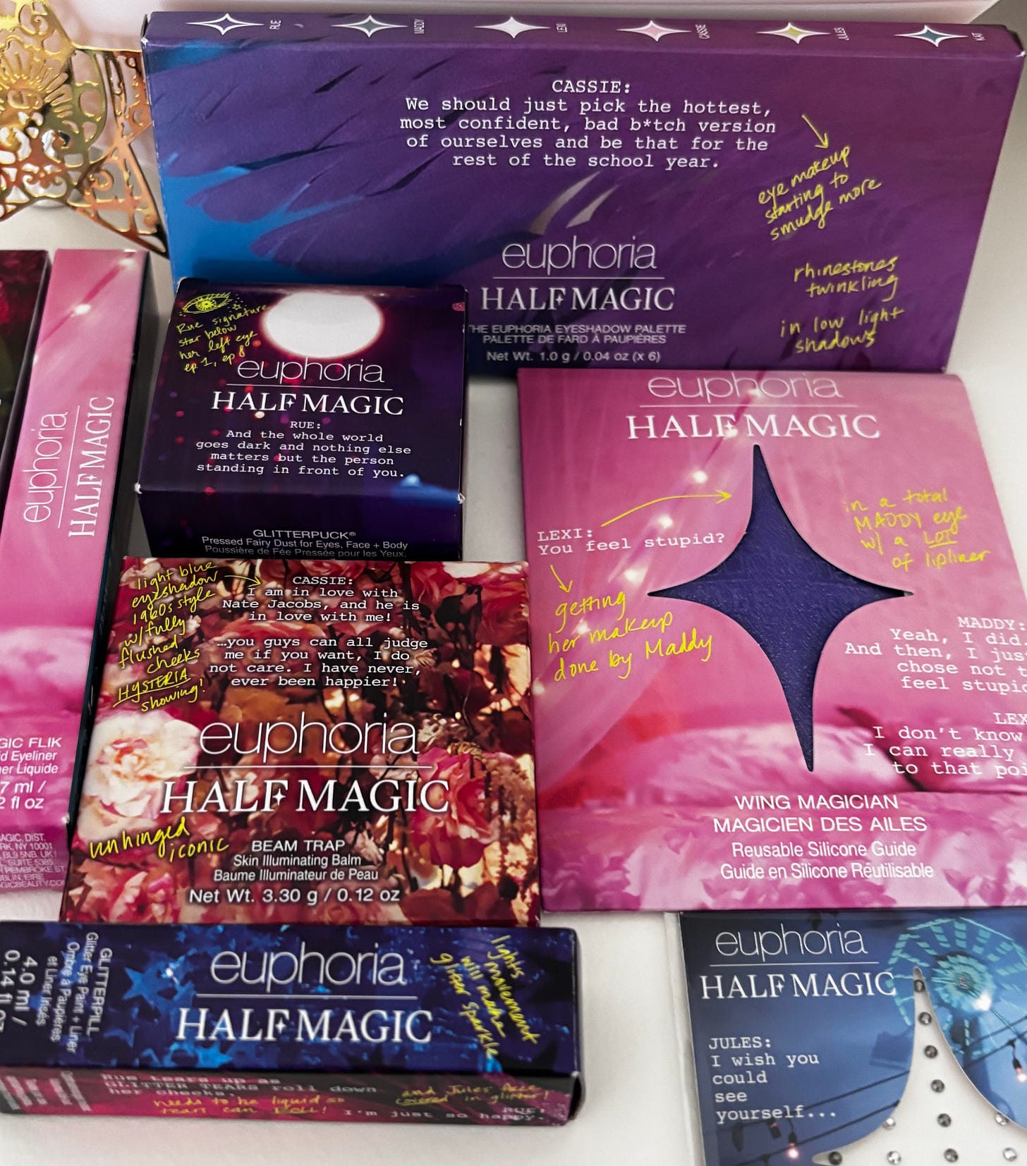 A flat-lay arrangement of the Half Magic x Euphoria collection, including the eyeshadow palette, Glitterpuck, lip gloss, and vibrant yellow applicator tool set against a backdrop of adhesive pearls.