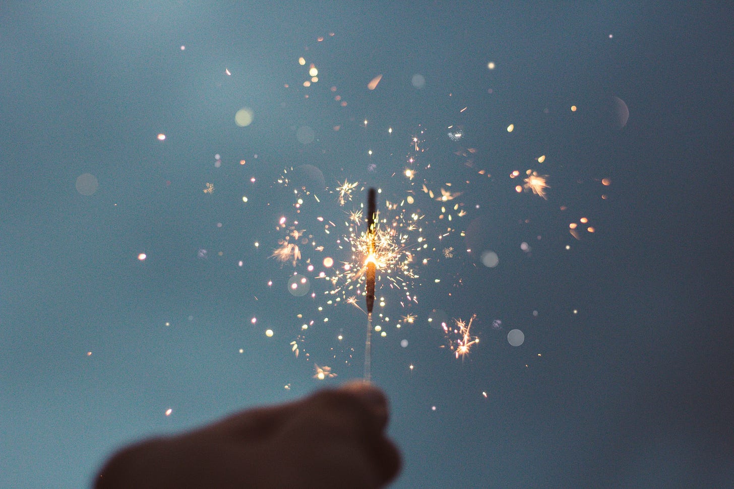 Sfondo opaco con tonalità azzurro piombo, mentre in primo piano compare sfuocata una mano che regge una fontanella scintillante di quelle che fanno piccoli fuochi d'artificio.
