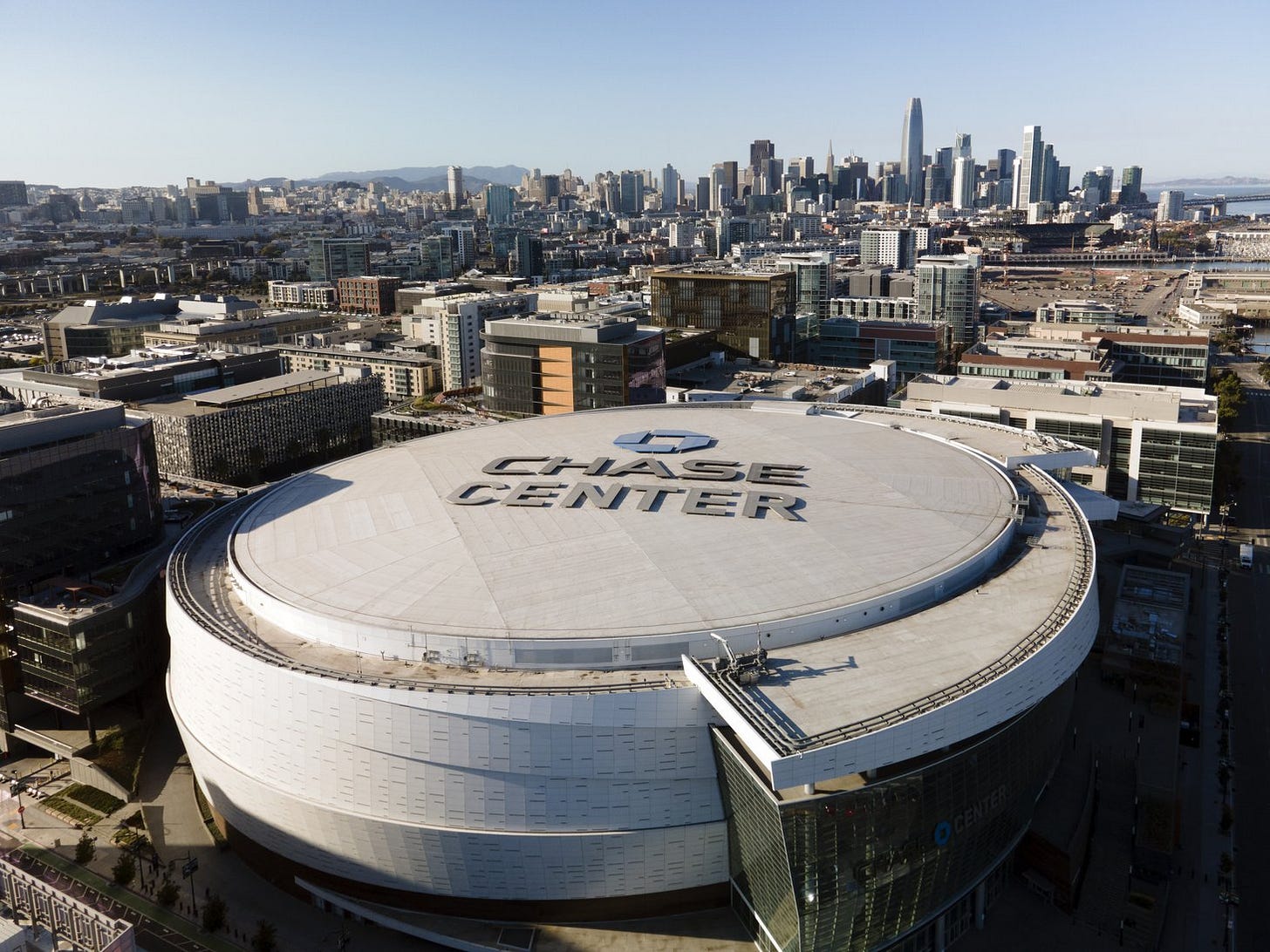 Aerial view of a round white arena with "Chase Center" written across its roof, with the San Francisco skyline behind it under a clear blue sky.