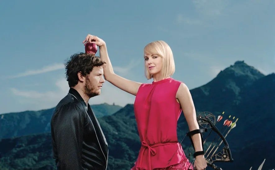 A white blonde woman in a pink dress holding a bow and arrows places an apple on the head of a white, brown-haired man with a beard wearing a robe