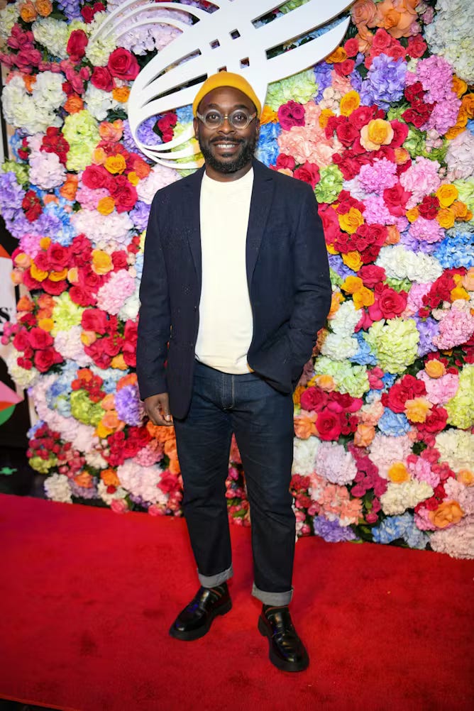 A man dressed in dress jacket, jeans and yellow beanie stands on a red carpet with a floral display behind him.