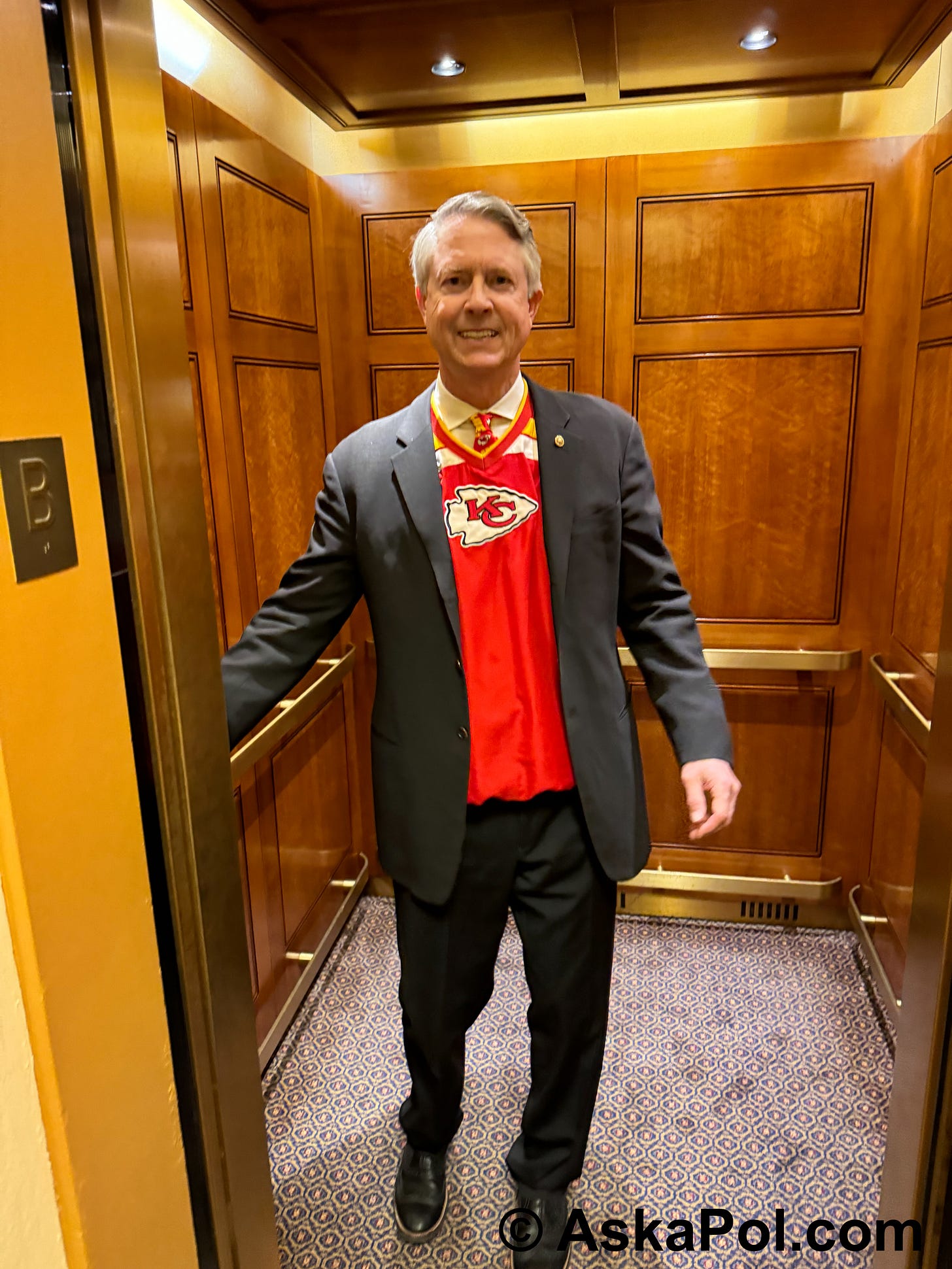 A man is smiling wearing a red Kansas City Chiefs jersey with suit coat on top as he hops elevator. Photo: Matt Laslo © www.askapolpolitics.com A man is smiling wearing a red Kansas City Chiefs jersey with suit coat on top as he hops elevator. Photo: Matt Laslo © www.askapolpolitics.com