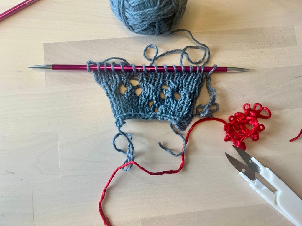 A knitted swatch on a needle with no waste yarn remaining, showing open live stitches at the cast-on edge.