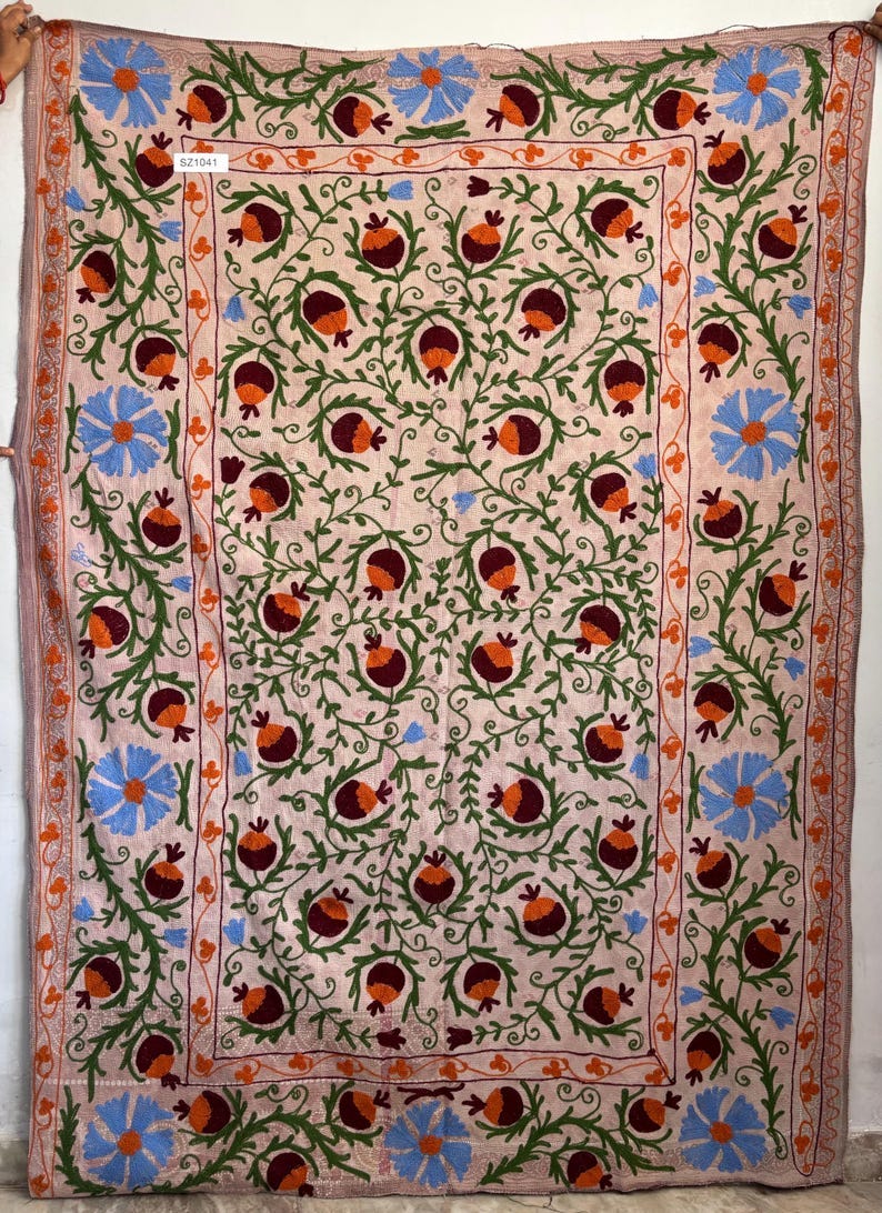May include: A pink embroidered fabric with a floral pattern. The fabric features a repeating pattern of green vines, orange and maroon pomegranates, and blue flowers. The fabric has a border of orange and green embroidery. The fabric has a tag that reads '521041'.