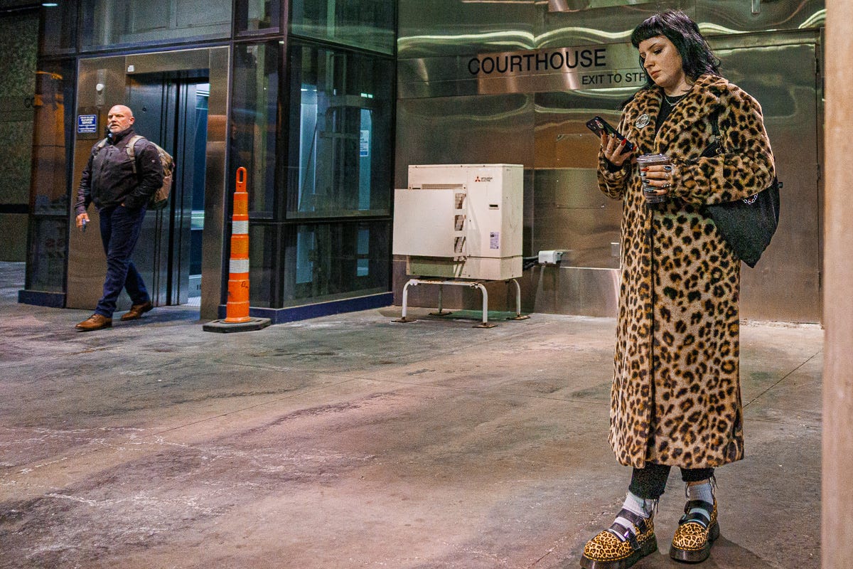 Woman in leopard print coat and shoes checking her phone at Courthouse Station in Boston, with a man exiting the glass elevator behind her. Woman in leopard print coat and shoes checking her phone at Courthouse Station in Boston, with a man exiting the glass elevator behind her.