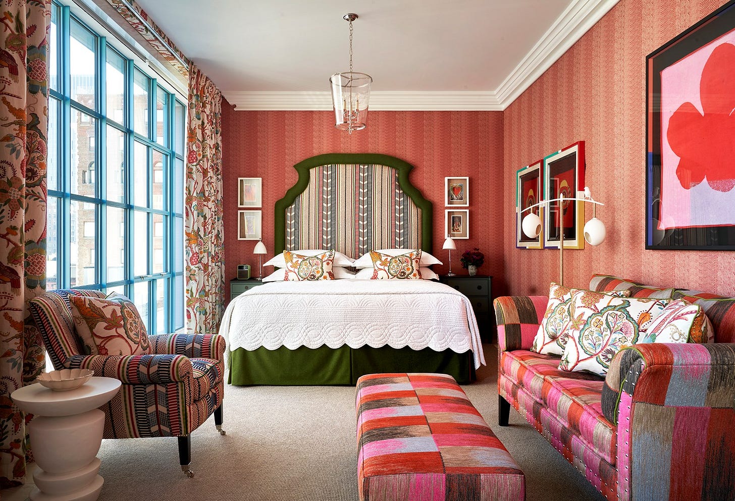 A dark green bed with a headboard upholstered in a muted striped fabric, a matching patterned arm chair on the right of the room with a white side table next to it. A Willow Ikat weave sofa and footstall on the left.