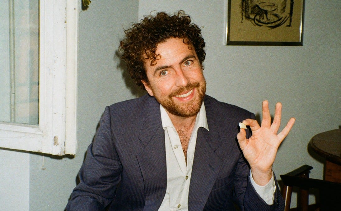 A photo of Michael McGovern sitting down. He is smiling, and appears to be pinching a small, undefined object between his index finger and thumb.