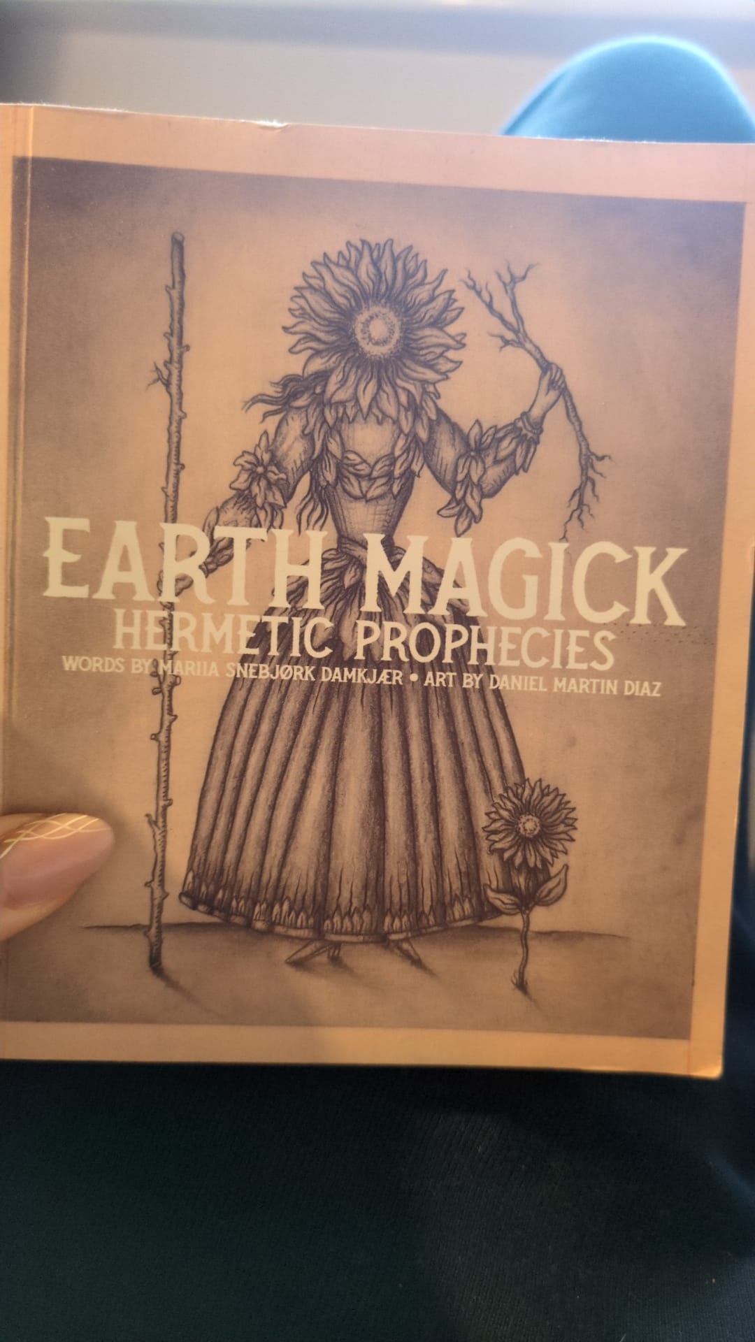 A photograph of the cover of a booklet titled “Earth Magick: Hermetic Prophecies.” The cover illustration shows a robed, human-like figure with a sunflower for a head, holding a staff in one hand and a branching twig in the other. A smaller sunflower grows at their feet. The artwork is rendered in sepia tones with fine line detail, giving it an occult, botanical feel.