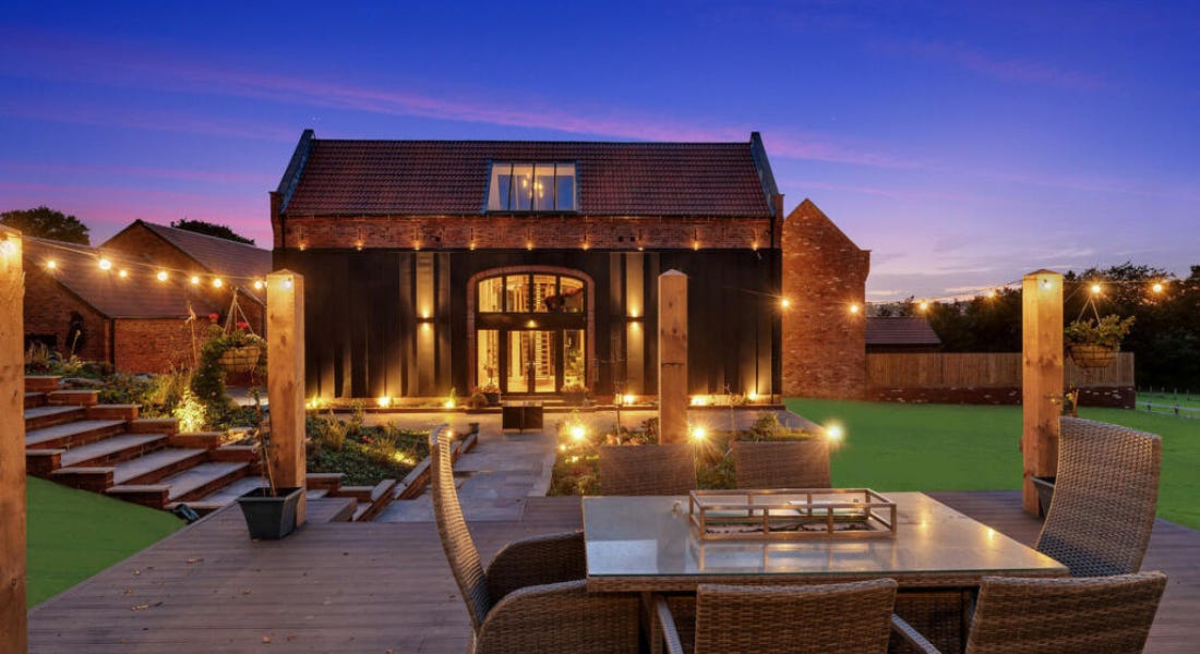 A large barn conversion house with decking and outdoor seating in front of it and a dark purple sky behind it