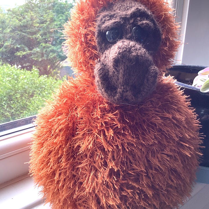 A knitted face applied to the smallest round, which is balanced on the largest round. Knitted in orange fluffy yarn for the rounds, and brown yarn for the face. 