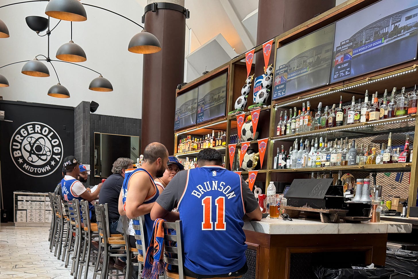 12:12 PMClaude responded: A crowded bar scene with patrons seated along a long bar counter, several wearing New York Knicks jerseys including one reading "Brunson 11.A crowded bar scene with patrons seated along a long bar counter, several wearing New York Knicks jerseys including one reading "Brunson 11." Multiple televisions above the bar display FIFA World Cup 2026 coverage. The Burgerology Midtown sign is visible on the back wall. Bottles of liquor line the shelves behind the bar, decorated with Knicks pennants and soccer balls. 12:12 PMClaude responded: A crowded bar scene with patrons seated along a long bar counter, several wearing New York Knicks jerseys including one reading "Brunson 11.A crowded bar scene with patrons seated along a long bar counter, several wearing New York Knicks jerseys including one reading "Brunson 11." Multiple televisions above the bar display FIFA World Cup 2026 coverage. The Burgerology Midtown sign is visible on the back wall. Bottles of liquor line the shelves behind the bar, decorated with Knicks pennants and soccer balls.