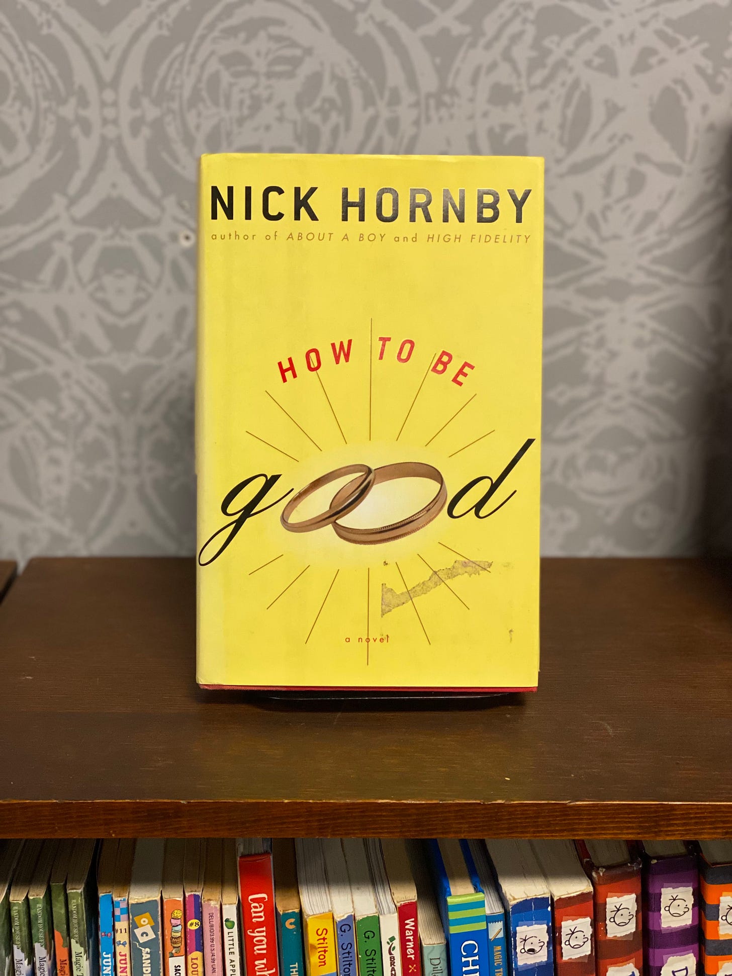 cover of Nick Hornby's How to Be Good, yellow background with type treatment and two wedding bands as the Os in "good"