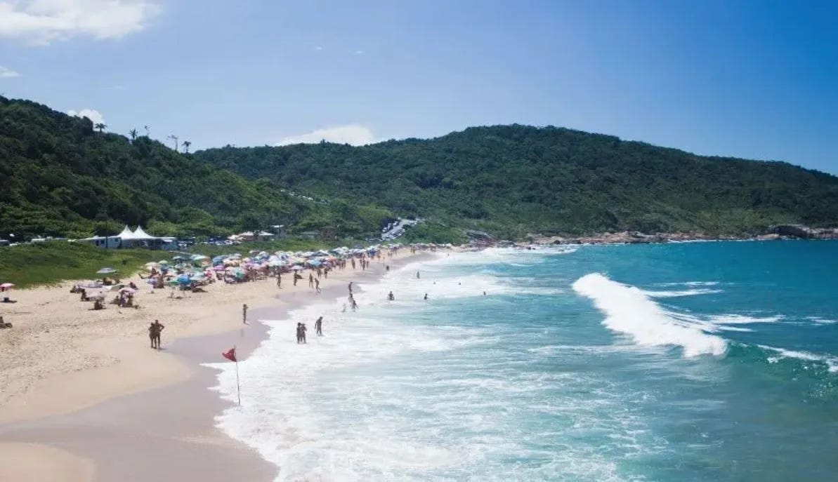 A wide sandy beach with turquoise waves and lush green hills in the background. Dozens of umbrellas line the shore, and people are seen swimming and walking along the surf. A red flag marks the shoreline near the center of the image.
