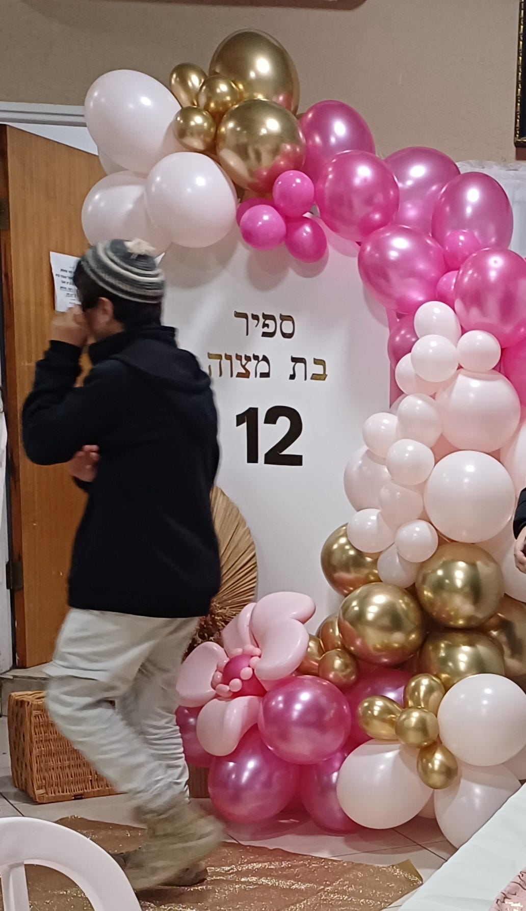 An elaborate white, pink and gold balloon decoration from the party, with a brother passing by during the party's setup.