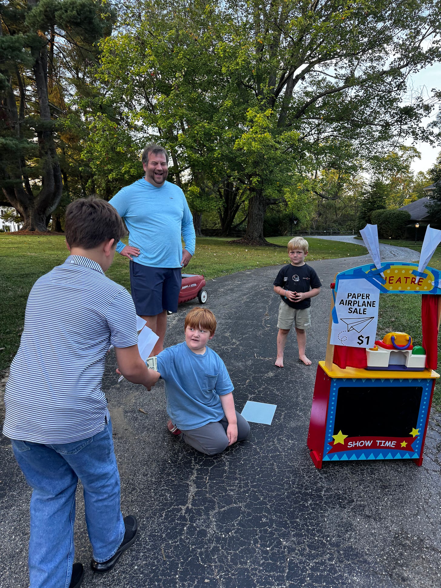 Boy sitting in his driveway folding paper airplanes on the spot, selling them for $1 each as a simple act of creativity and entrepreneurship.
