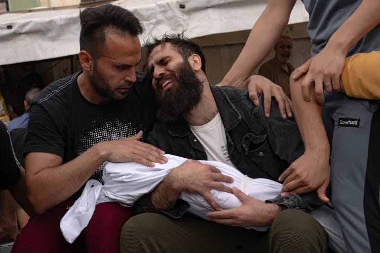 a palestinian man crying while he holds the body of a child in a shrowd and another palestinian man has his arm around him and is visibly trying to console him