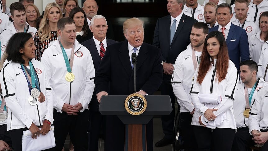 Trump speaks at a lectern flanked by men and women in suits or white tracksuits, many with medals around their necks. Trump speaks at a lectern flanked by men and women in suits or white tracksuits, many with medals around their necks.