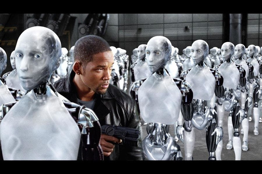 Um monte de robôs com rosto humanoide, todos da mesma cor (branca e cinza), estão einfileirados horizonaltamente e verticalmente. Entre a primeira e segunda fileira da perspectiva do observador, passa Will Smith, um homem negro de cabelo bem curto, com uma jaqueta de couro e uma arma na mão. O robô mais próximo da câmera, logo a frente de Smith, está olhando rpa ele de canto de olho. Smith não parece ter notado.