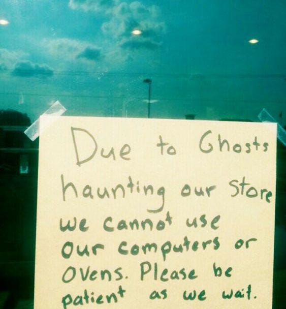Blue/gray skies with clouds in the background and a yellow post it note taped to the foreground window reading "due to ghosts haunting our store, we cannot use our computers or ovens. Please be patient as we wait"