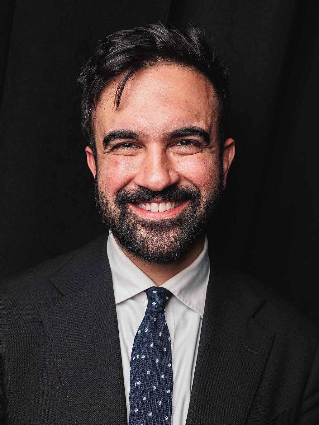 Mamdani with mussed hair and beard with a blue tie with white spots smiling against a black background Mamdani with mussed hair and beard with a blue tie with white spots smiling against a black background