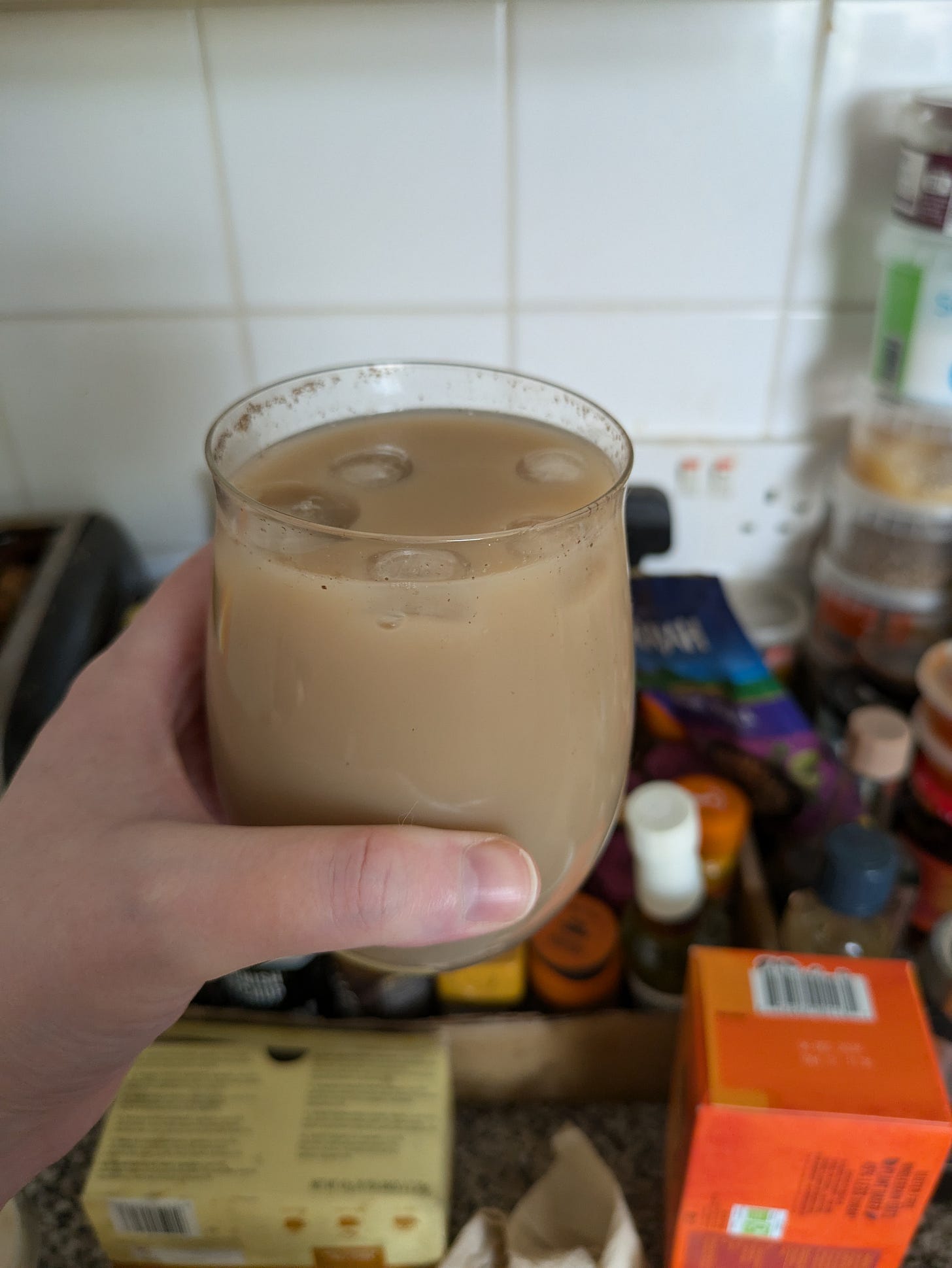 ID. For variety I took this photo of my hand holding a nice glass full of iced chai in my kitchen. In the blurry background there is a box of spices and condiments, the corner of a toaster and a stack of questionably filled pots of seasonings. ID. For variety I took this photo of my hand holding a nice glass full of iced chai in my kitchen. In the blurry background there is a box of spices and condiments, the corner of a toaster and a stack of questionably filled pots of seasonings.