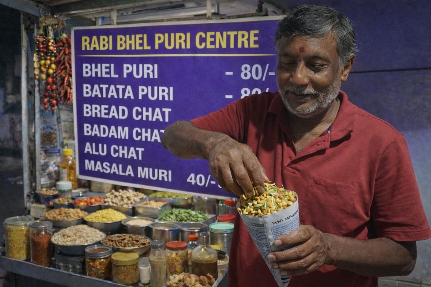 A man in red tee shirt preparing Indian snacks in a paper cone at a streetside stall with a price chart visible in the background. A man in red tee shirt preparing Indian snacks in a paper cone at a streetside stall with a price chart visible in the background.