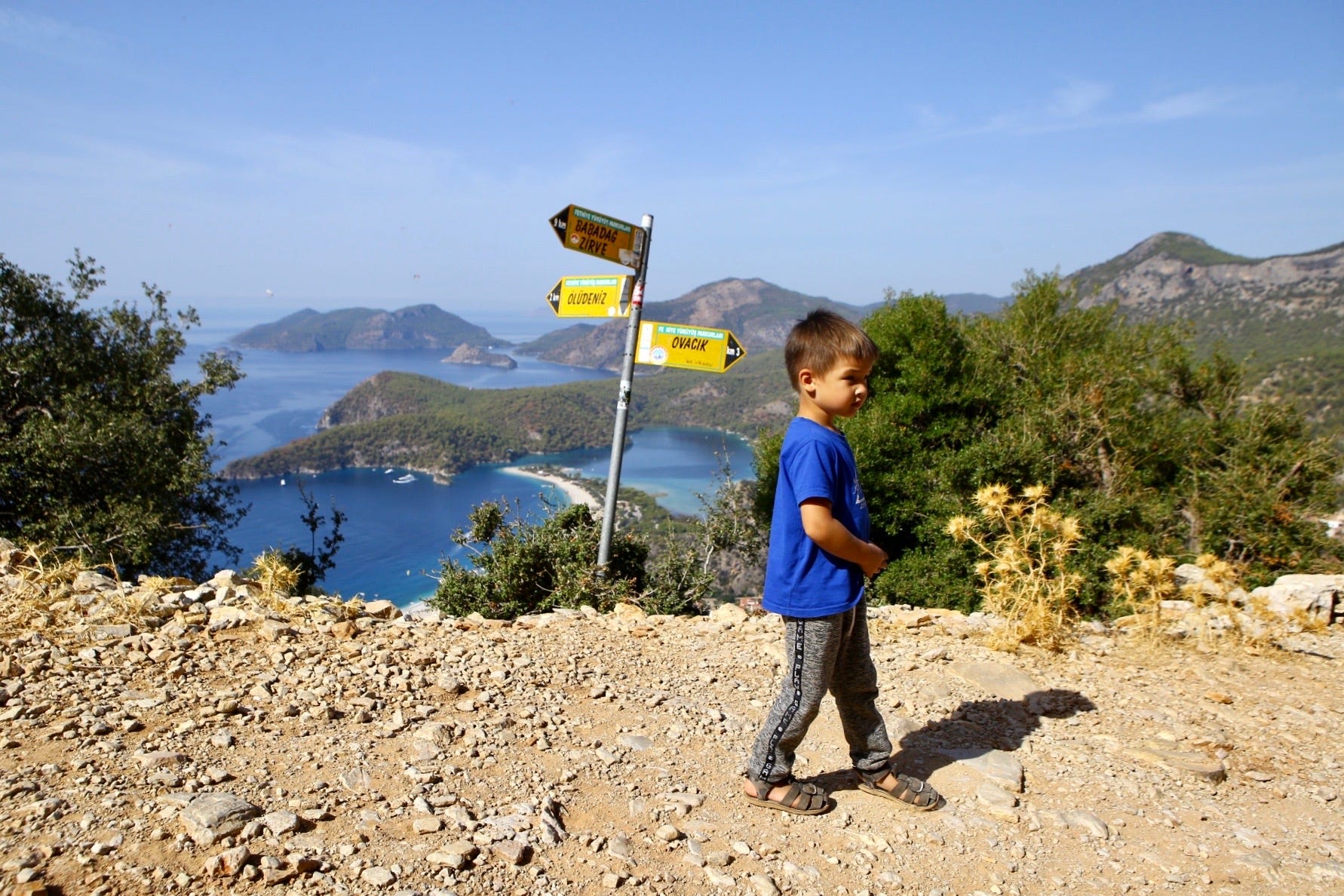 A boy in a blue shirt stands in front of directional signs for Turkish towns above the sea and a beach.