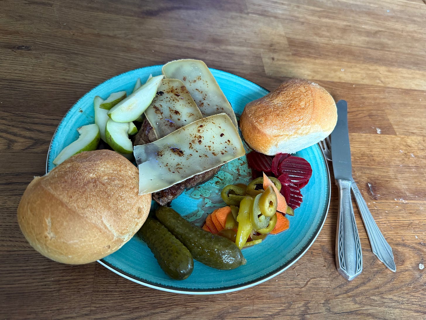 Image of a blue plate with two rolls, a large piece of steak covered in cheese with the juices melting the top, on the sides are pear slices, pickles and pickled vegetables. Next to the plate is one fork and one knife. 