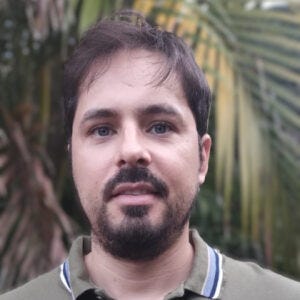 Fotografia de um homem de barba curta e cabelo castanho, vestindo uma camisa polo verde com detalhes azul e branco na gola. Ele está ao ar livre, com um fundo desfocado de folhas verdes, possivelmente de palmeiras, sugerindo um ambiente natural. Sua expressão é neutra, e a iluminação suave destaca seu rosto de forma equilibrada.