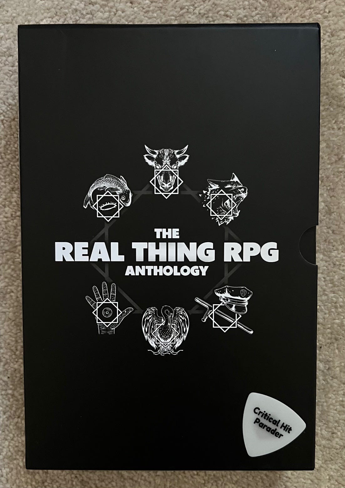 Black slipcase for The Real Thing RPG Anthology. Six white symbols are depicted, which represent images from Faith No More albums and songs. A white Critical Hit Parader guitar picks lies on the bottom right corner of the box like a woodpecker from mars.