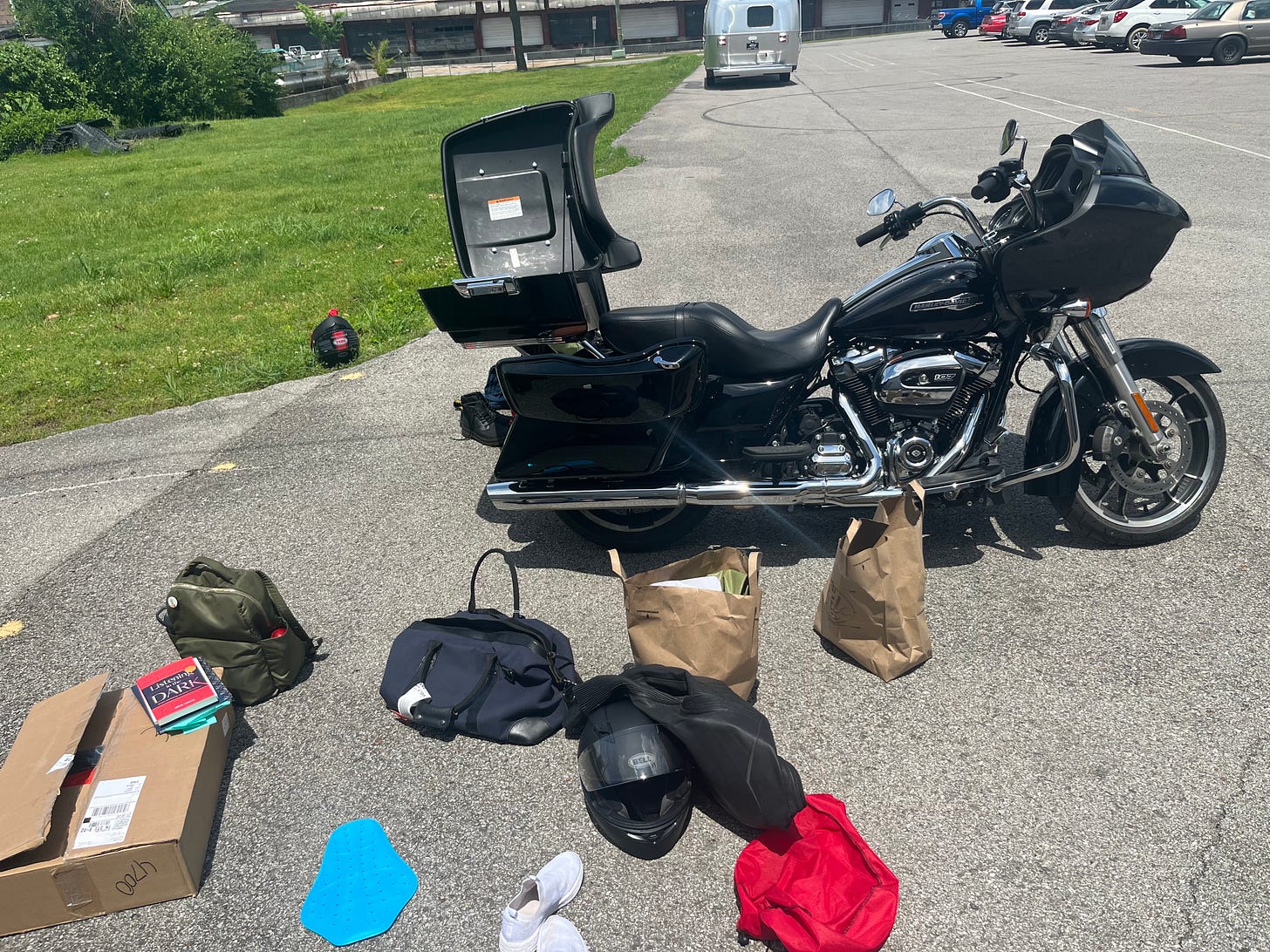 A parked motorcycle with its storage section and pockets open. Boxes, books, and bags are strewn around on the ground waiting to be packed into the bike.