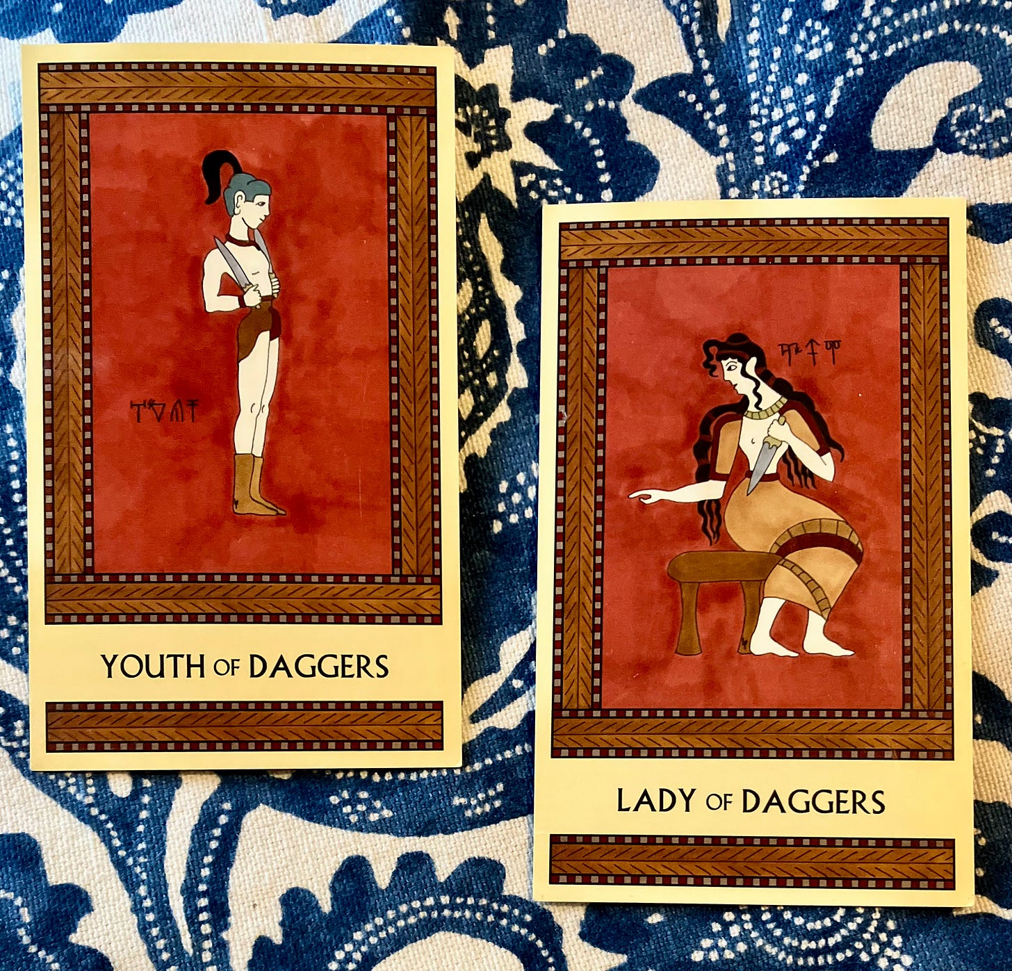 Two Minoan Tarot cards side by side on a blue-and-cream print background. Both cards are painted in shades of deep red and brown. The Youth of Daggers shows a Minoan teenage boy wearing a loincloth and short boots, standing and facing right. He holds a dagger in each hand, his arms bent at the elbows so the daggers rest on his shoulders. The Lady of Daggers depicts a Minoan woman seated on a stool, facing right but with her head turned to the left. Her right hand points to the left. In her left hand she holds a dagger pointed downward. 