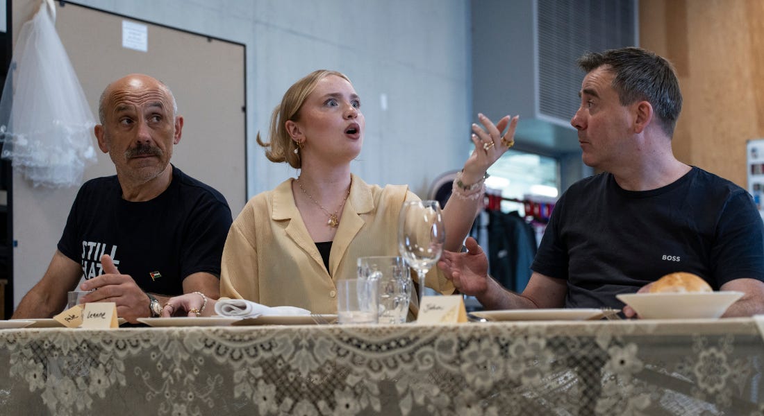 Three people sit behind a long table. A young women in the middle raises her left arm and speaks to someone off camera, looking alarmed. The man to her right has a questioning look on his face, whilst the man to her left is looking at her in surprise. Three people sit behind a long table. A young women in the middle raises her left arm and speaks to someone off camera, looking alarmed. The man to her right has a questioning look on his face, whilst the man to her left is looking at her in surprise.