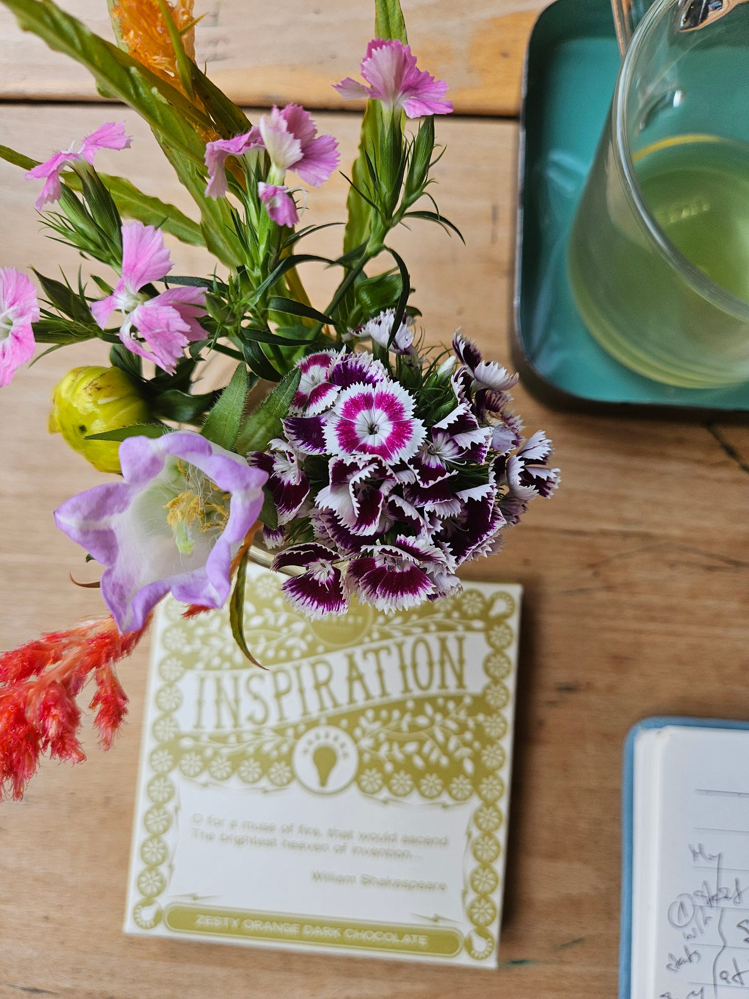 The image shows a decorative arrangement on a wooden surface. There is a bouquet of flowers, including purple and white flowers with a fringed edge, and a light purple flower with a yellow center. In the center of the image, there is a package of CocoPzazz's Inspiration Dark Chocolate. The packaging is white with a gold pattern and the word "INSPIRATION" prominently displayed in the centre. To the right, there is a glimpse of a green drink in a glass. The image shows a decorative arrangement on a wooden surface. There is a bouquet of flowers, including purple and white flowers with a fringed edge, and a light purple flower with a yellow center. In the center of the image, there is a package of CocoPzazz's Inspiration Dark Chocolate. The packaging is white with a gold pattern and the word "INSPIRATION" prominently displayed in the centre. To the right, there is a glimpse of a green drink in a glass.